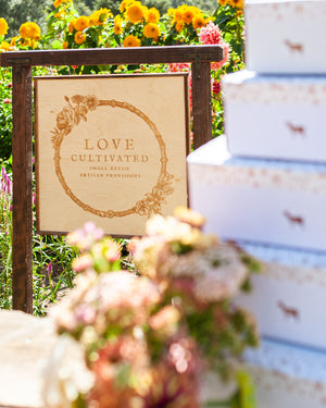 A picture of the Love Cultivated sign, with gift boxes in the foreground wrapped with custom tape and a goat stamp. Appear next to a bouquet of flowers.